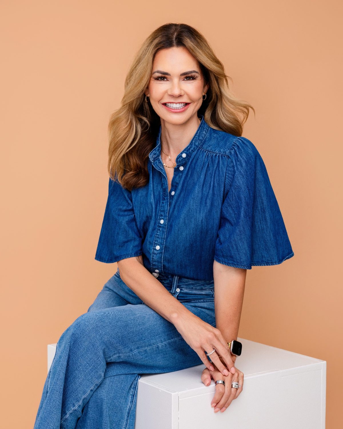 Alison Lumbatis in a denim top and jeans, seated on a white cube against a warm peach background.